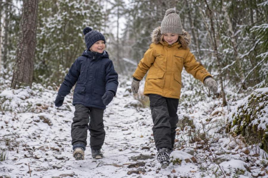 To børn i mørkeblå og okkerfarvede vinterjakker på en let snedækket skovsti.
